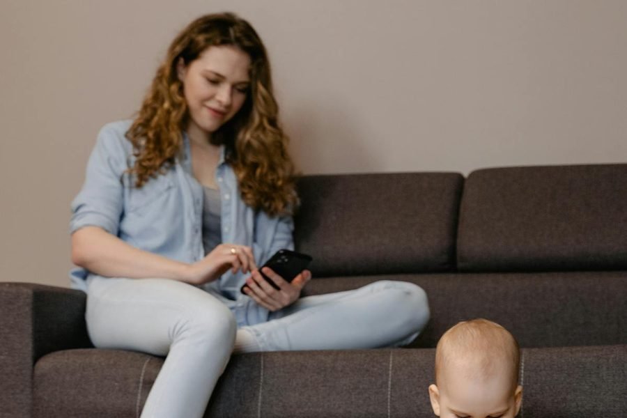 Une maman détendue remplit le formulaire sur son téléphone, assise sur le canapé pendant que son bébé joue à côté.