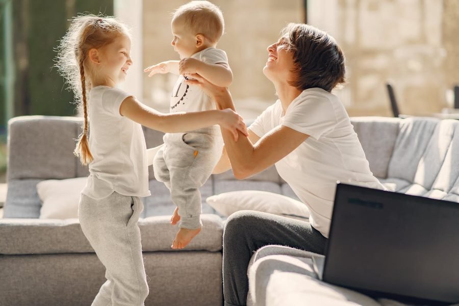Deux enfants jouent et rient avec leur babysitter dans le salon, lumière chaleureuse de fin d'après-midi.