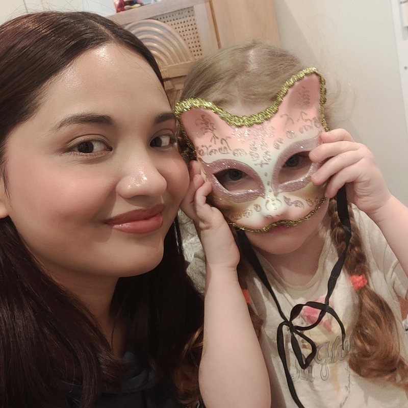 Une babysitter Bloom Buddies souriante joue avec une petite fille qui tient un masque de chat vénitien devant son visage — moment complice du quotidien.